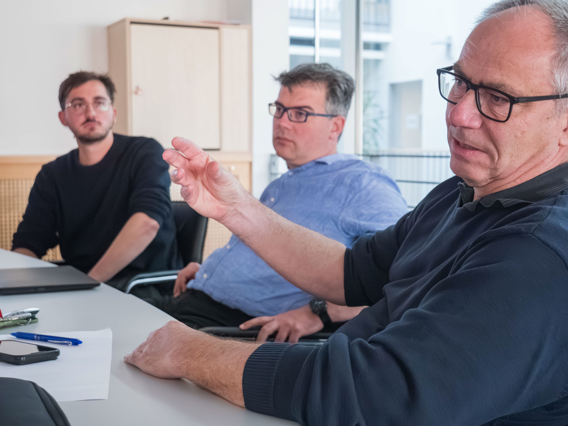 Ein Mann gestikuliert im Vordergrund. Im Hintergrund sitzen zwei weitere Männer